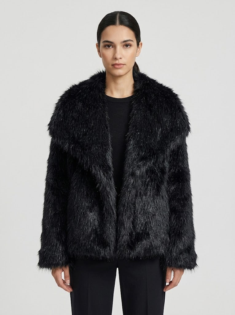 A model wearing a black faux fur jacket with a wide collar over a black top and black pants, photographed against a light gray studio background.
