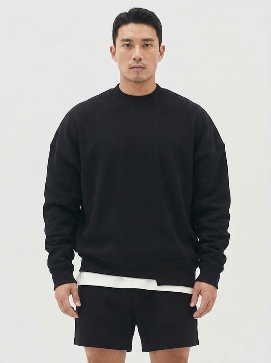 A man wearing a black crewneck sweatshirt with a white underlayer peeking out at the hem, paired with black shorts, against a plain light gray studio background.